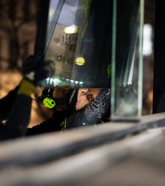 Närbild av tre Bollan-tekniker som koncentrerat installerar ett glasparti på kvällen med stadens ljus i bakgrunden.